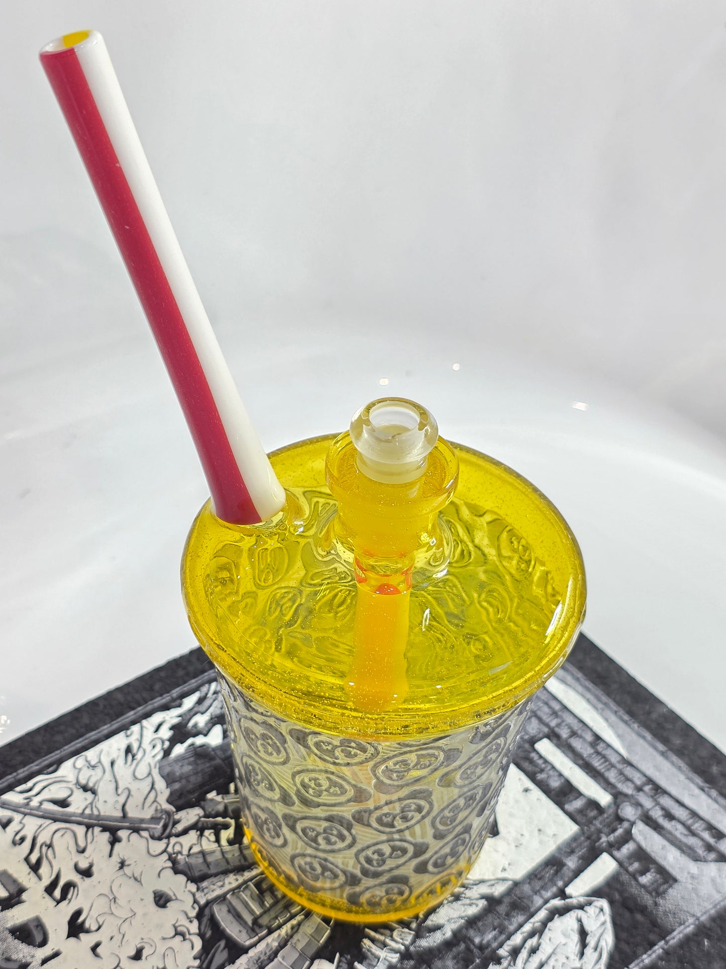 Slop Glass x Disk Glass Yellow glass rig cup with patterned base, red straw, and decorative lid on a reflective surface.